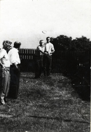 Photograph of two men standing at the left of the photograph, watching two other men who are in the centre of the picture, one of whom has just thrown a quoit, which can be seen in the air; behind the men is a fence and to the right are bushes; one of the men to the left is bare-chested; they have been identified as members of the British Legion Team