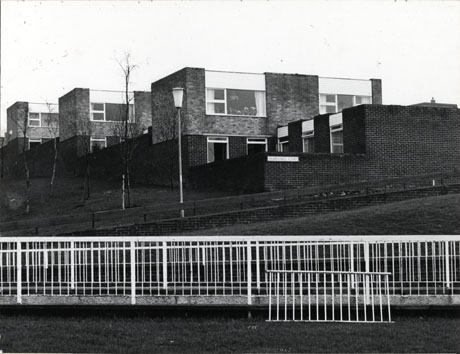Photograph of three blocks of two-storeyed houses one behind the other; in front of the blocks is a wall on which the name Brandling Court can be seen; in front of the Court are a grassy slope and fences along a path