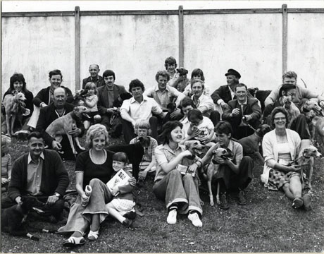Photograph of a group of approximately nineteen people and six children sitting in front of a fence on grass; eleven dogs can be seen with the group; the group has been described as Whippet Racing Crowd