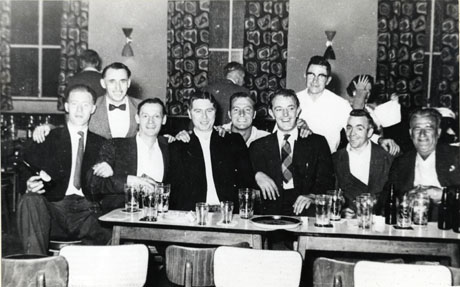 Photograph of six men sitting at two Formica-topped tables pushed together; behind them are three other men looking at the camera; behind the group three other people looking elsewhere can be seen; there are glasses on the table and three windows with curtains and two wall-lights can be seen behind the group; the photograph has been described as Peterlee Workingmen's Club Members, 1950