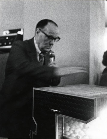 Photograph of a middle-aged man wearing a suit and tie and spectacles; he is holding a microphone in his left hand and gesturing with his right, which is blurred; he is sitting at a machine in which balls can be seen; he has beeb identified as Walter Parker (Housey Caller)