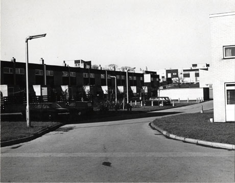 Photograph of a block of flat-roofed two-storey houses with a fence at the rear and cars parked in the road; the block is on the left of the picture; in the centre of the photograph is the surface of the road, which curves to the right of the picture; on the right are the exteriors of further blocks of houses; the photograph has been identified as being of Peterlee