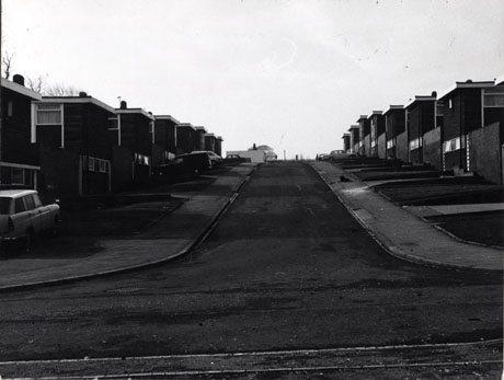 Photograph of a road running uphill away from the camera; on either side of the road are pavements, open-plan grass plots and two-storey houses with flat roofs; six cars are parked in front of the houses; the road has been identified as Dart Road, Peterlee