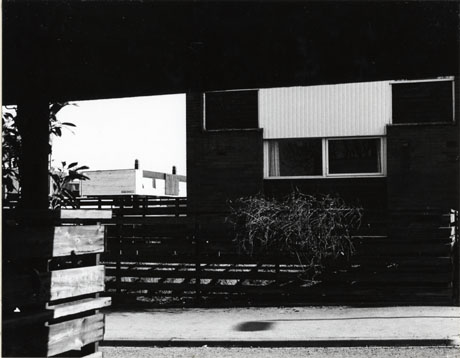 Photograph of the exterior of the ground floor of a house, with, beyond it, the top storey and flat roof of another house; the photograph has been taken through an opening in another building; it has been identified as Dart Road, Peterlee