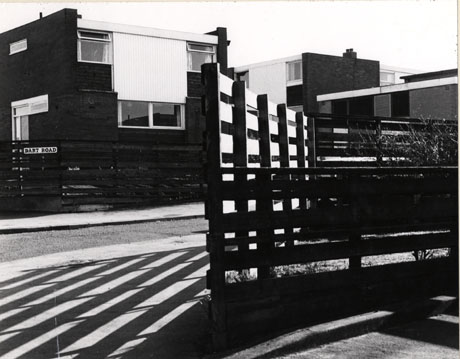 Photograph of a two-storey house with a fence in front on which a street sign reading Dart Road [Peterlee] can be seen; at the front of the photograph is wooden fence which is casting bars of shadow on the pavement