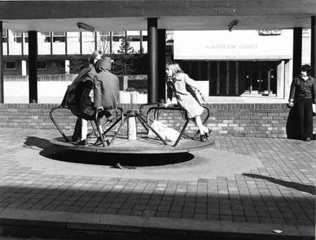 Photograph of a children's roundabout on which there are three girls playing; they are wearing coats and the tree in the distance is leafless; behind the roundabout is the underside of the roof of a walkway or a similar structure; behind the roof is the facade of the Magistrates' Court; a boy can be seen standing on the right of the picture; the photograph has been identified as Children's Roundabout, Peterlee Town Centre, 1977