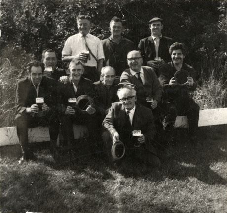 Photograph of ten men sitting on a plank of wood, drinking glasses of beer and holding quoits in their hands; they are posed against trees and grass; they have been identified as British Legion Quoits Team, Peterlee