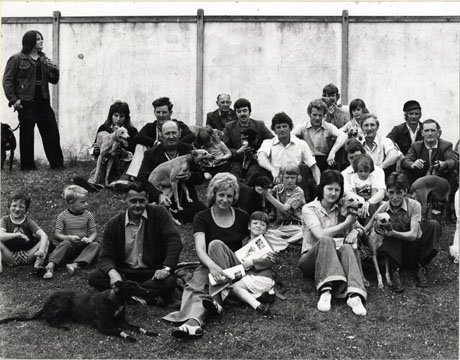 Photograph of groups of people in front of a wall, sitting on the ground, with whippets, as in pete0022 and pete0035; the crowd has been identified as being in Peterlee