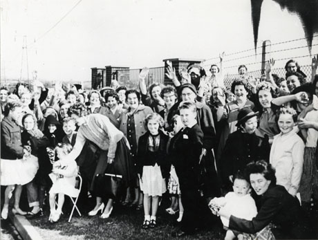 Photograph of a crowd of women and children in front of a fence and gateway; the kerb of a pavement can be seen at the left of the picture; it has been identified as occurring on the occasion of the visit Queen Elizabeth II and Prince Philip to Peterlee