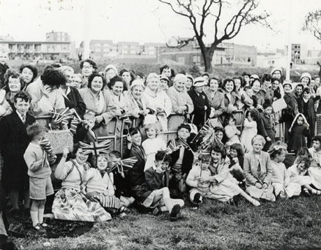 Photograph of a crowd of women, standing behind metal barriers, with children sitting on the ground in front of the barriers; behind the crowd is open ground and, behind that, houses and larger buildings in the distance; the photograph has been identified as occurring on the occasion of the visit to Peterlee of Queen Elizabeth II and Prince Philip