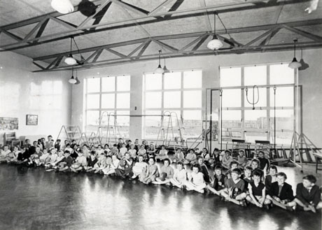 Photograph of the interior of a gymnasium at a school, identified as Eden Hall School, Peterlee; the ceiling, two large windows, equipment, pictures on the wall and the polished floor of the building can be seen; a group of approximately seventy children aged approximately between seven and ten years are sitting cross-legged on the floor in front of the windows; children on the front row are holding musical instruments