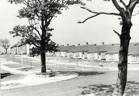 Photograph showing the facades of a row of bungalows built up the side of a gentle hillside; at the top of the hill are four semi-detached houses; in front of the bungalows are gardens, a road, patches of grass and saplings; immediately in the foreground are two trees set in the pavement, one of which is at the junction of the road running down the hill with a road running a right angles with it; the photograph has been identified as Junction at Kirkland Road, Peterlee