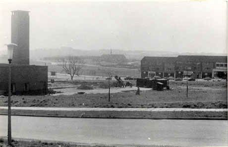 Photograph showing a view over the site of Peterlee, with a fire station on the left; in the middle of the picture, a building with a small spire, possibly a church, can be seen in the distance; on the right of the picture, a parade of shops can be seen in the distance; in the foreground, are what appears to be work in progress with, possibly, work huts, turned earth and machinery; the photograph has been described as View From Surtees Road, Peterlee