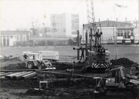 Photograph of an excavation with a mechanical digger on the right of the picture; in the centre is the base of a mobile crane; on the right is another digger and a sign reading Ogden; planks and other debris are lying across the excavation which stretches the width of the photograph at the front; in the distance a low building on the left, a tall building in the centre, and a low building on the right can be seen; the photograph has been identified as Fine Fare Supermarket Under Construction at Peterlee