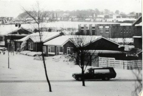 Photograph of Peterlee under snow; in the foreground are saplings and a road running from right to left of the picture on which a car is travelling; behind the car are bungalows and, behind them, a flat-roofed building and further buildings in the distance