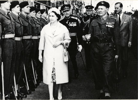 Photograph showing Queen Elizabeth II wearing a coat and hat inspecting soldiers who are standing to attention on the left of the photograph; a soldier, possibly of the Parachute Regiment, is accompanying the Queen; behind them the Lord Lieutenant of County Durham, another soldier, and Prince Philip, can be seen; the photograph has been described as recording a visit of the Queen to Peterlee