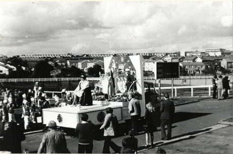 Photograph of a float on which there are a young woman sitting on a throne, two children behind her dressed in costume standing before a coat of arms, and the number 25; indistinct groups of people, including members of a jazz band, can be seen either side of the road looking at the float; in the distance, houses can be seen; the photograph has been identified as depicting the Carnival to mark the 25th Anniversary of Peterlee