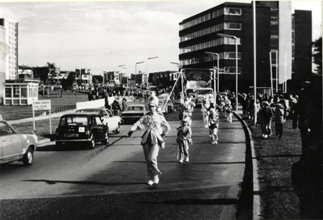 Photograph of a road running towards the camera on which there are a procession of children in the uniform of a jazz band, followed by a lorry carrying a float; on the opposite side of the road, travelling away from the camera, is a line of cars; crowds of people can be seen in the distance on either side of the road; a tall block of offices or flats is on the right of the picture; the photograph has been identified as recording the 25th Anniversary Carnival in Peterlee