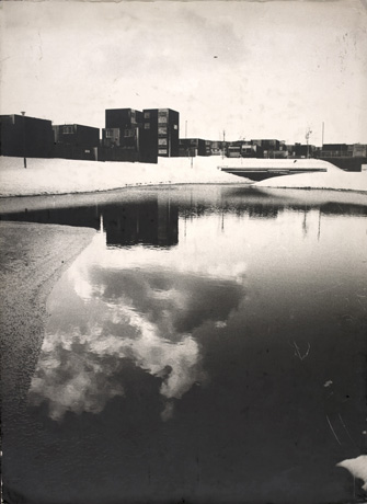 Photograph showing, in the foreground, an extensive pool of water in which clouds are reflected; beyond the pool are tall blocks of buildings, and low flat-roofed buildings, on the horizon, with snow-covered slopes between them and the pool, which has been identified as Sunny Blunts Pool, Peterlee