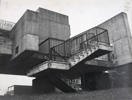 Photograph, taken from below, showing two flights of concrete steps, with steel railings, attached to a building consisting of two concrete blocks, raised off the ground by further, smaller concrete blocks; a concrete wall is at the top of the steps and a concrete bridge appears to lead from the two concrete blocks to the left; the photograph has been described as Steps Going Over Bridge Over Sunny Blunt Pool, Peterlee