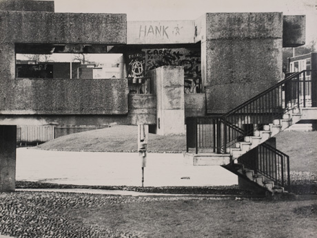 Photograph showing, as in pete0083, concrete steps with steel railings, at the right of the picture, leading up to the end of a concrete structure, only the first two blocks of which can be seen in pete0083; the structure can now be seen as a long bridge extending over a pool of water, which can be seen behind the bridge; there are graffiti on the concrete, including the word, Hank, and the symbol of the Campaign For Nuclear Disarmament; the photograph has been described as Front View of Steps Over Sunny Blunts Pool, Peterlee