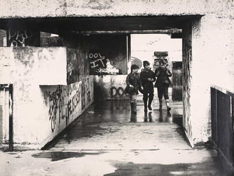 Photograph showing three boys, aged approximately between seven and nine years, walking over a concrete floor under a flat concrete roof between concrete blocks, which are covered in graffiti; they have been described as Children Walking Over Sunny Blunts Bridge, Peterlee