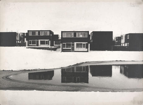 Photograph showing a pool of water extending from the right of the picture with snow-covered ground surrounding it; in the background are the fronts of two flat-roofed houses overlooking the pool and the side of three other rows of flat-roofed houses; the photograph has been described as Houses Looking Onto Sunny Blunts Pool, Peterlee