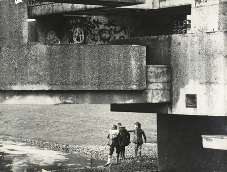 Photograph of three boys, aged approximately between seven and nine years, standing with their backs to the camera and with water at their feet and grass beyond them; above them, forming most of the picture, are the large concrete blocks, which are part of the bridge over the pool; graffiti can be seen on one of the blocks; the photograph has been described as View of Sunny Blunts Bridge