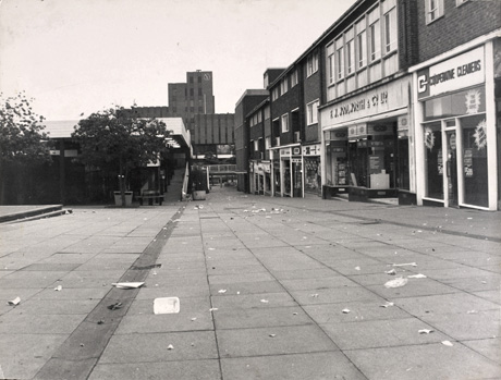 Photograph looking along a paved area with shops on the right, including Co-operative Cleaners and Woolworth, leading to a stair on the left and a tall concrete building, with a clock on its tower, in the distance; the area is the shopping centre in Peterlee and the building in the distance is Lee house, Headquarters of the Peterlee Development Corporation; the shopping centre is deserted and there is litter strewn everywhere near the shops