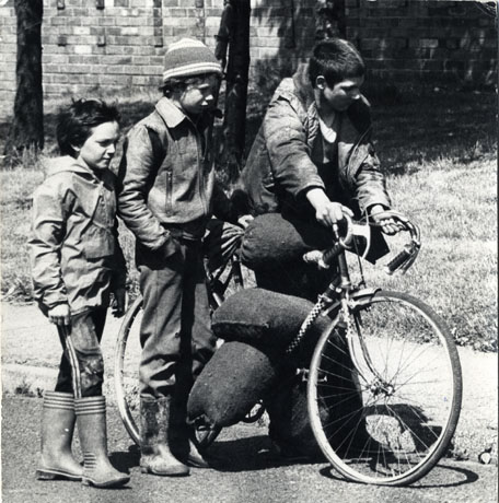 Photograph of three boys, aged between ten and fourteen years, walking along a street with part of a brick wall and a strip of grass behind them; the largest of the boys is wheeling a bicycle on the crossbar of which three full sacks are balanced; the boys are dressed in Wellington boots, trousers and anoraks; the photograph has been identified as Young Boys Carrying Coal on Bike, Peterlee