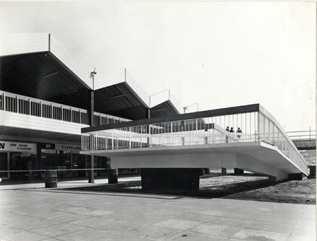 Photograph showing the underside and back of a ramp and its railings; behind the ramp, part of the facades of shops and the gables of the building above them can be seen indistinctly; the ramp has been identified as being in Peterlee Town Centre