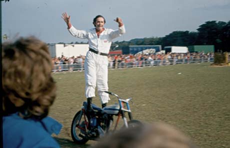 Photograph of a man in white overalls standing on a small motor bicycle while it is in motion; crowds are watching him from behind barriers at the edge of the field on which he is performing; he has been identified as performing at Peterlee Carnival