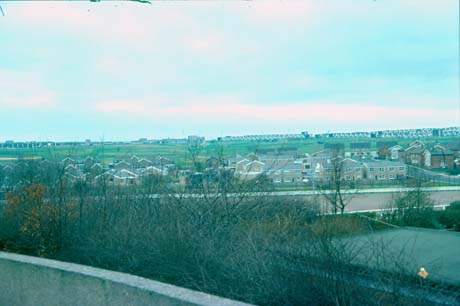 View, taken from above, showing flat-roofed houses on the horizon and houses with pitched roofs in the middle distance; immediately in front of the camera are the bare branches of trees and the roof of a building; the picture has been identified as a view of an area near the town centre in Peterlee