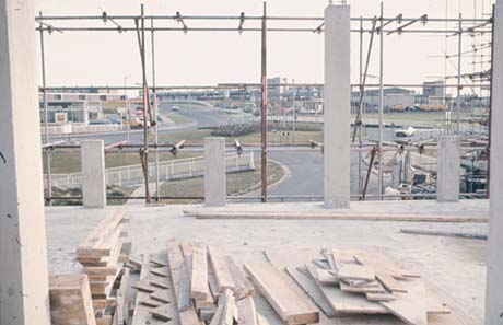 Photograph showing piles of wooden planks lying on a concrete floor with scaffolding at its edge through which a view of a roundabout and buildings beyond it can be seen; it has been identified as a photograph taken of the view from Lee House, Peterlee, as it was being built