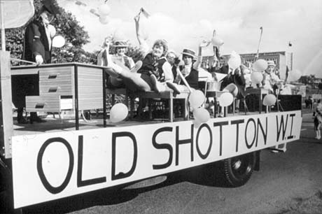 Photograph showing the side of a lorry with the words Old Shotton Womens' Institute along it; on the lorry are eight women dressed as schoolgirls and a school teacher; they are sitting at desks and are wearing gym slips and boaters; a drum majorette can be seen in the road behind the float; the float is proceeding along a road with a building behind it which appears to be in Peterlee
