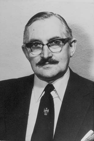 Photograph of the head and shoulders of a man wearing a suit and tie, which has a coat of arms on it; he is wearing glasses and a moustache and has been identified as Councillor Cliff Heslop