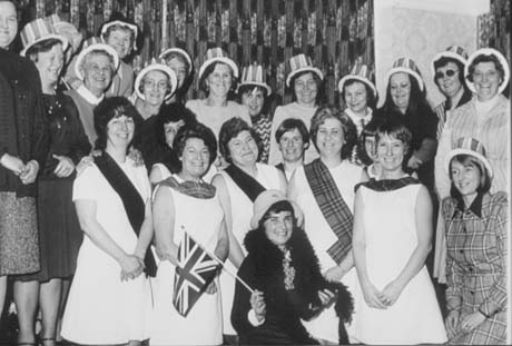 Photograph of fifteen women, wearing dresses and skirts and striped hats, standing in front of the wall of a room round eight women wearing light-coloured dresses and tartan stoles; a further woman is in front of them wearing a feather boa, cloche hat and holding a Union Jack; they have been identified as members of old Shotton Womens' Institute