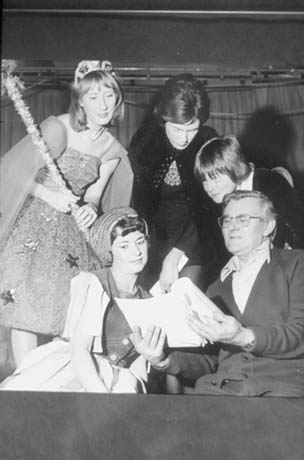 Photograph showing a woman in a tutu, carrying a wand and wearing a tiara, standing behind another woman, who is wearing sixteenth-century-looking costume of dress and headdress and talking to a man in a cardigan, who is holding a script; behind the man are two other women, who are also looking at the script; they have ben identified as members of old Shotton Womens' Institute