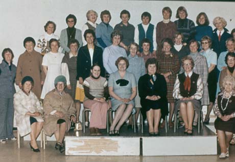 Photograph of thirty one women posed in three rows against the wall of a room; they are wearing coats, dresses, trouser suits, pinafore dresses, and four are sitting on a platform of two boxes; they have been identified as members of old Shotton Womens' Institute