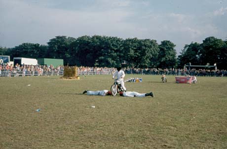 Photograph showing a man wearing white overalls riding a motor cycle between two men wearing white overalls lying on the ground in a field with spectators surrounding it; there are other obstacles, such as a bale of straw and a ramp, in the field; the motor cycle demonstration has been identified as being part of Peterlee Carnival