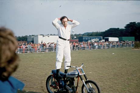 Photograph showing a man in white overalls standing on a motor bicycle with his hands behind his head; he is performing on a field with crowds watching from behind barriers at the perimeter; he has been identified as taking part in Peterlee Carnival