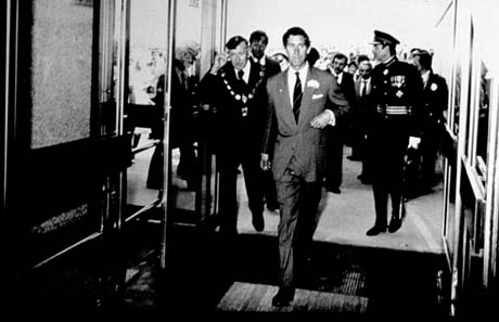 Photograph showing The Prince of Wales walking through the doorway of a building towards the camera; he is wearing a suit and tie and is carrying a garment over his arm; behind him are a man in a chain of office and the Lord Lieutenant in uniform; other people are behind them; the occasion has been identified as the opening of Peterlee Leisure Centre