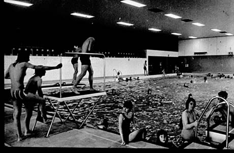 Photograph of an indoor swimming pool with people swimming in the pool and three people on the diving board; a woman is sitting on the side of the pool and another is about to climb out; the pool has been identified as that in Peterlee Leisure Centre