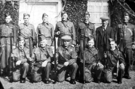 Photograph showing twelve men in uniform and one man in civilian clothes, posed against the exterior of a window and door of a house; they are wearing battle dress and are carrying weapons; one man is wearing battle dress and a cap; they have been identified as members of the Old Shotton Local Defence Volunteers