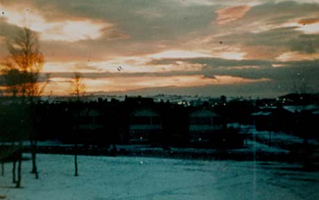 Photograph taken at sunset showing the colours of the sky and a dark mass of buildings with the occasional light; in the foreground are a slope and trees; the photograph has been identified as Church Close, Peterlee