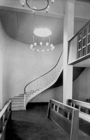 Photograph showing a staircase with metal balusters curving round to a gallery with wooden balusters; above the staircase is a round chandelier and the backs of pews are on the right of the picture; the staircase has been identified as leading to the organ gallery in St. Cuthbert's Church, Peterlee