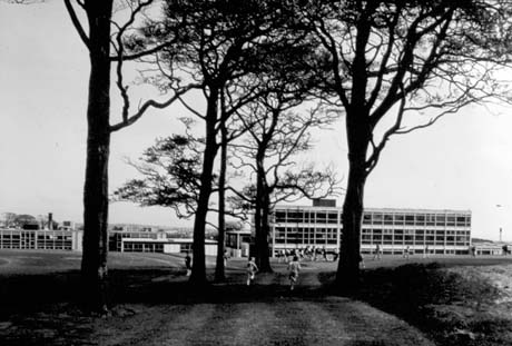 Photograph of the exterior of five blocks of buildings consisting of windows and concrete pillars separating the windows; the blocks are one, two and three storeys high; they have been identified as Shotton Hall School, Peterlee; indistinct figures can be seen near them