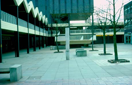 Photograph showing a raised walkway at the left and front, the back of a ramp, a paved floor, the side of a block with a shop and saplings, identified as part of The Town centre, Peterlee