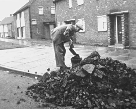 Photograph of a man, wearing a suit and cap, bending over a pile of coal in the gutter in front of a brick terraced house; the man has a shovel in his hand with which he is putting coal into a bucket;as the pavement is shining, it can be assumed that rain is falling or has recently fallen; the scene has been identified as being in Peterlee