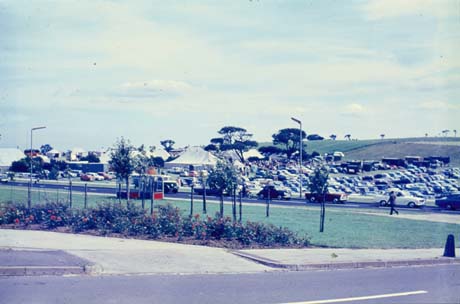 Photograph, taken from across a road and a piece of grass verge, of tents, surrounded by rows of parked cars; hillside can be seen behind the tents and cars; the scene has been identified as Show Ground, Peterlee